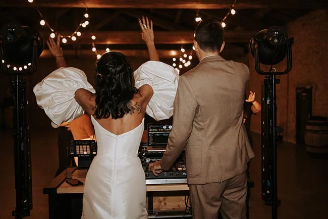 Couple de mariés de dos dansant devant un DJ sous des guirlandes lumineuses dans une ambiance festive et tamisée.