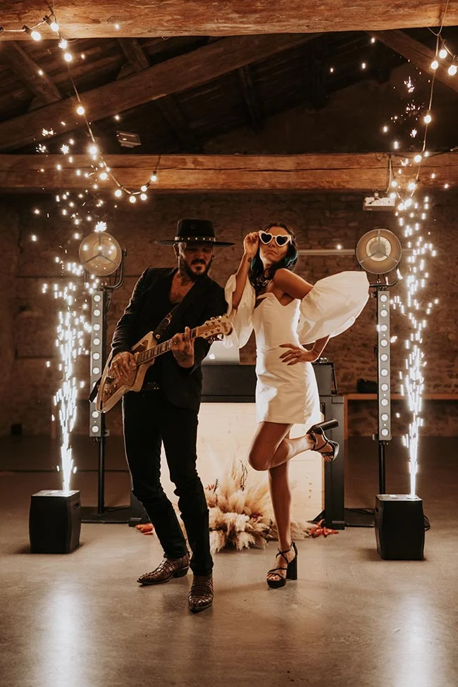 Couple stylé et excentrique posant avec une guitare sous des étincelles et des guirlandes lumineuses dans une grange.