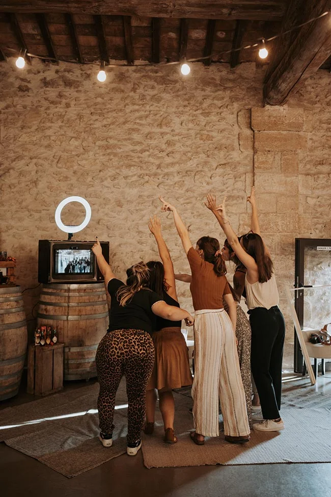 Groupe d'amies s'amusant devant un photobooth vintage dans une grange rustique aux tons chauds.