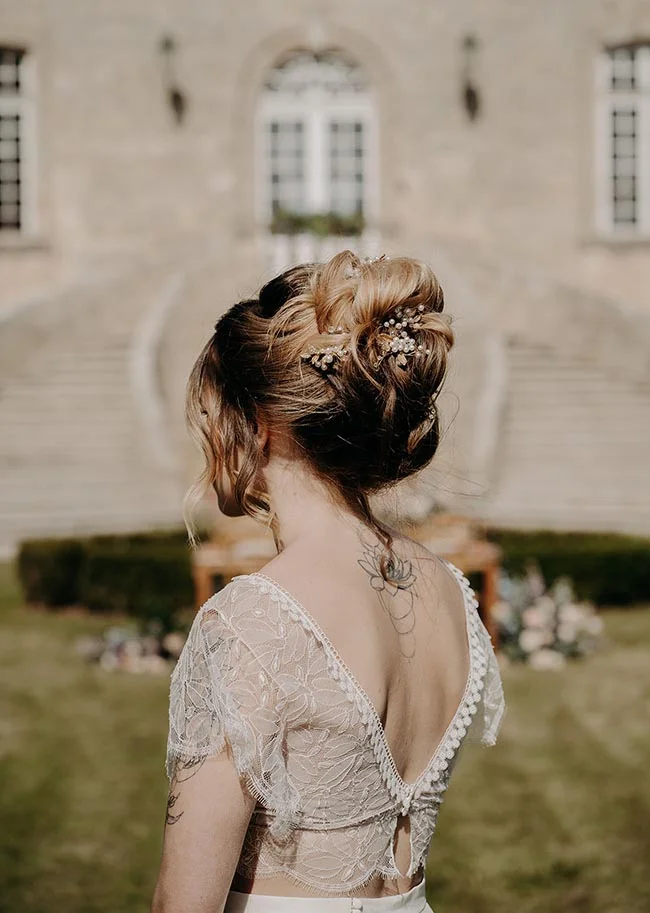 Dos d'une mariée en robe de dentelle blanche, coiffure relevée ornée de bijoux, devant un manoir romantique.