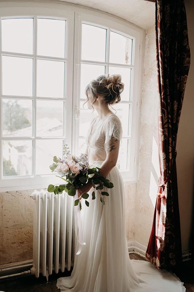 Mariée de profil en robe blanche et bouquet pastel, baignée de lumière près d'une fenêtre dans une ambiance romantique.