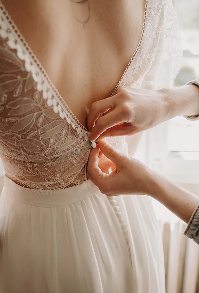 Gros plan sur les mains boutonnant le dos en dentelle blanche d'une robe de mariée romantique.
