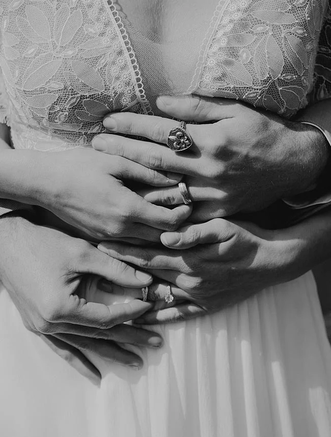 Gros plan en noir et blanc des mains d'un couple marié entrelacées, mettant en valeur leurs alliances et bijoux.