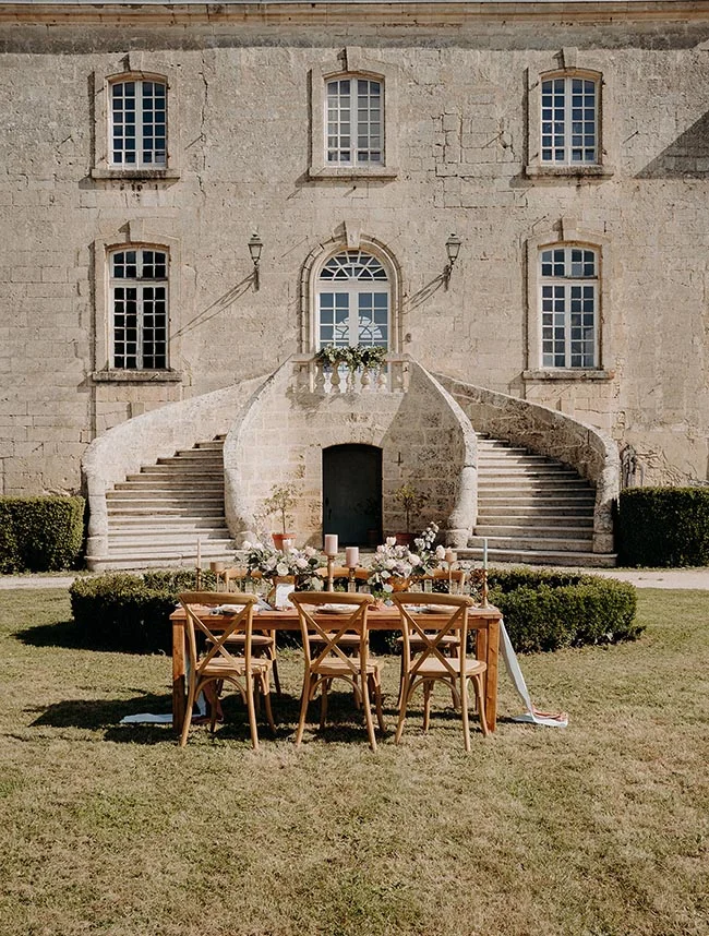 Table de réception élégante devant un château en pierre avec un escalier monumental, ambiance romantique et champêtre.