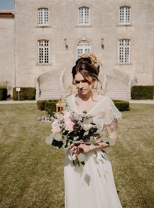 Mariée en robe blanche tenant un bouquet pastel devant un château classique, ambiance romantique et lumineuse.