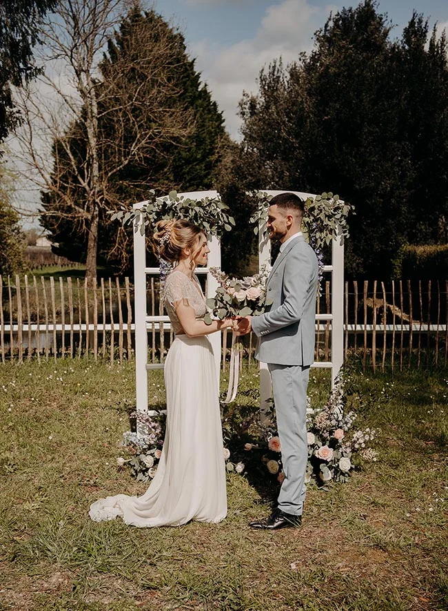 Jeunes mariés s'échangeant des vœux sous une arche fleurie dans un jardin champêtre, ambiance romantique et lumineuse.