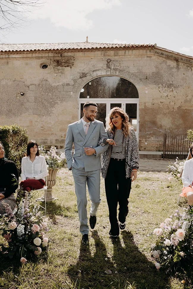 Un couple élégant marche dans un jardin devant un vieux bâtiment en pierre lors d'un mariage romantique.