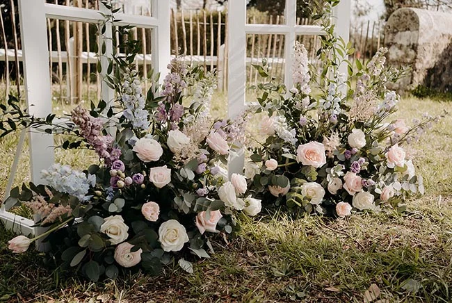 Compositions florales romantiques aux tons pastel au pied d'une arche blanche dans un jardin.