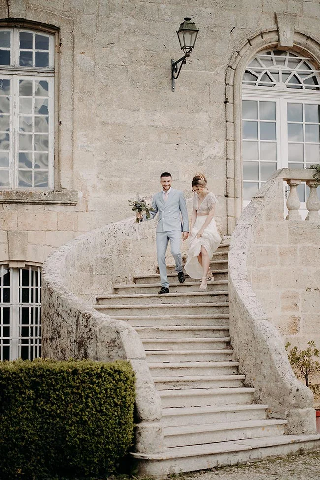 Jeunes mariés élégants descendant un escalier en pierre devant un château, ambiance romantique et lumineuse.