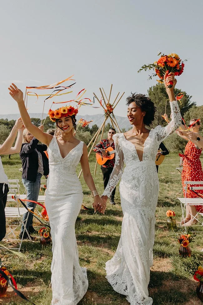 Deux mariées rayonnantes en robes blanches et couronne de fleurs célèbrent leur union lors d'un mariage folk en plein air.