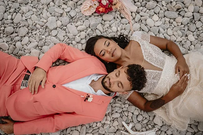 Jeunes mariés allongés sur des galets à Nice, elle en robe blanche et lui en costume corail, dans une ambiance romantique.