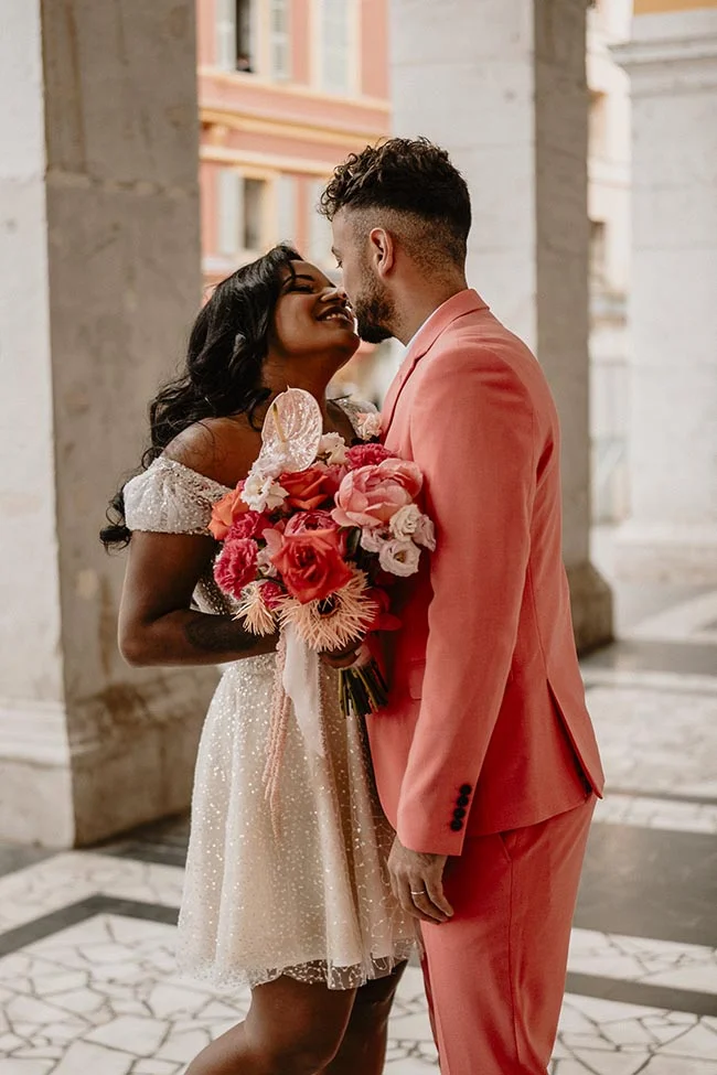Couple souriant s'embrassant, elle en robe blanche courte et lui en costume corail, avec un bouquet coloré à Nice.