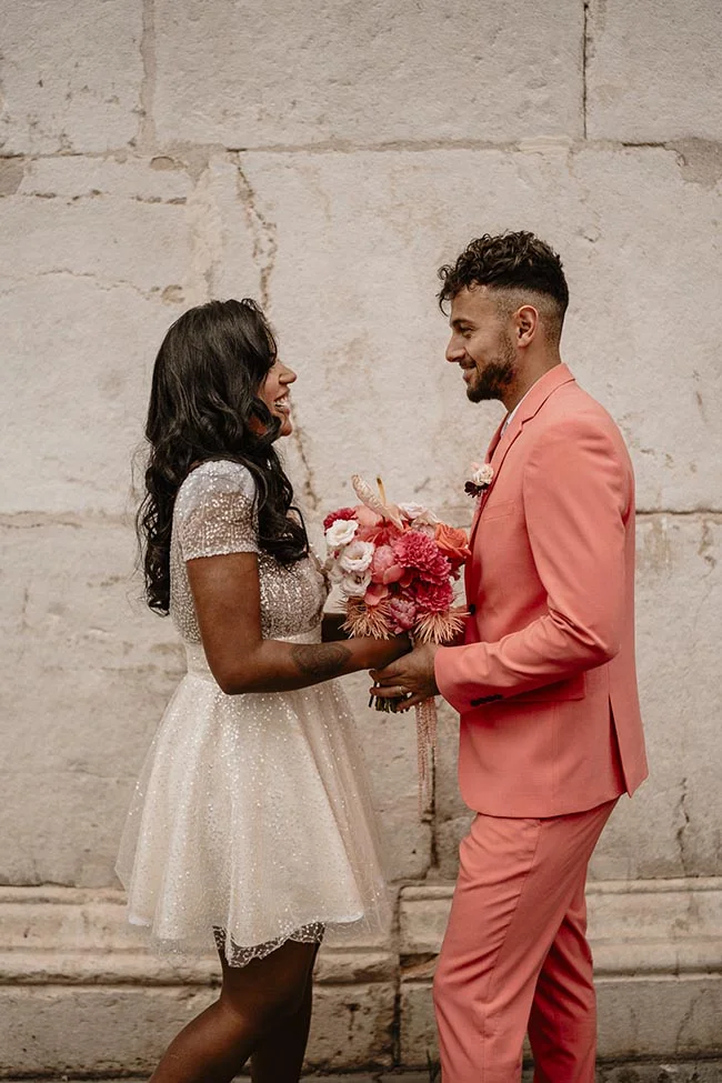 Couple souriant lors d'un elopement, elle en robe courte scintillante et lui en costume corail avec un bouquet coloré.