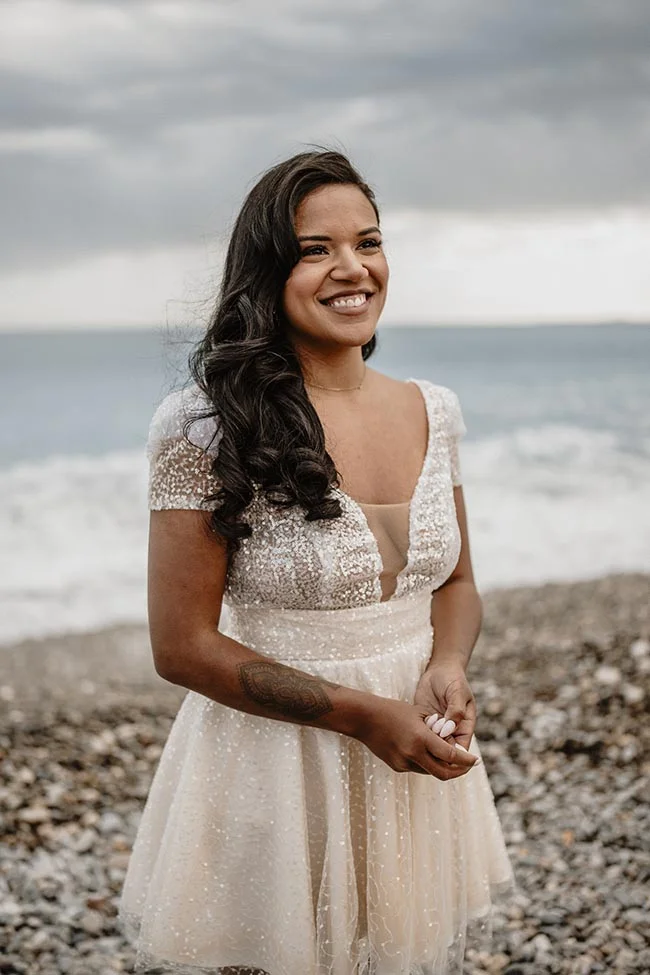 Une mariée souriante en robe blanche scintillante pose sur une plage de galets à Nice sous un ciel nuageux.