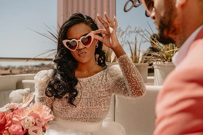 Une mariée espiègle en robe scintillante portant des lunettes en forme de cœur lors d'un elopement ensoleillé.