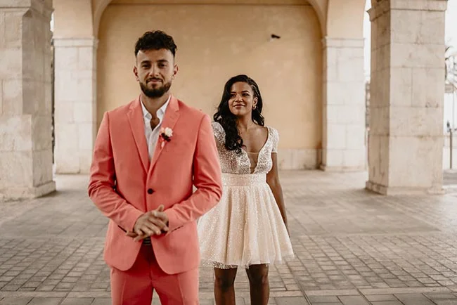Couple souriant pour un elopement à Nice, l'homme en costume corail et la femme en robe blanche courte et scintillante.