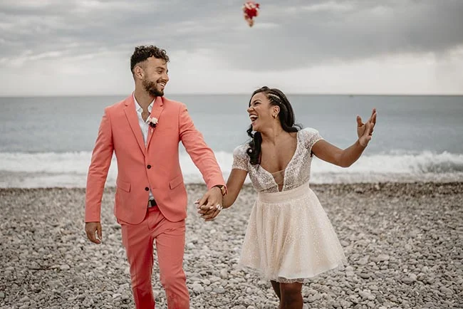 Couple riant main dans la main sur une plage de galets, lui en costume corail et elle en robe blanche courte.