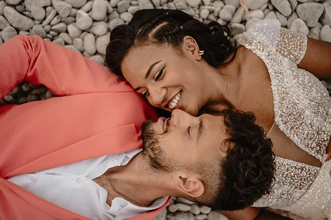 Couple marié souriant, allongé sur des galets, portant un costume corail et une robe blanche scintillante.