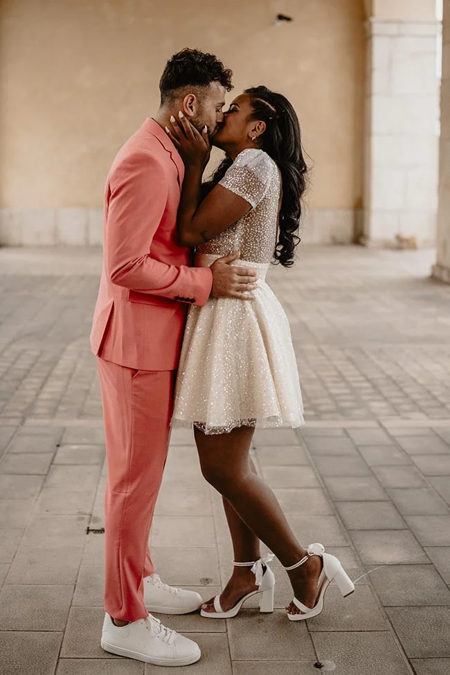 Couple s'embrassant avec passion, elle en robe blanche courte et lui en costume corail, dans un cadre urbain épuré.