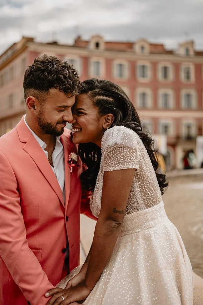 Couple complice et souriant lors d'un elopement, l'homme en costume corail et la femme en robe blanche scintillante.
