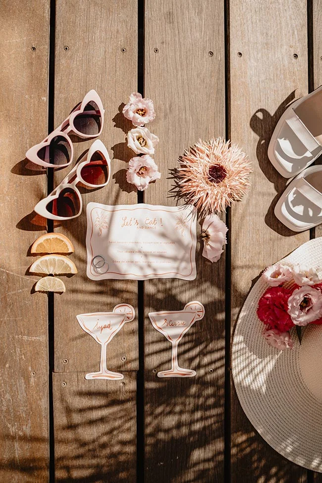 Accessoires de mariage estivaux et papeterie rose et blanche disposés sur un deck en bois ensoleillé.