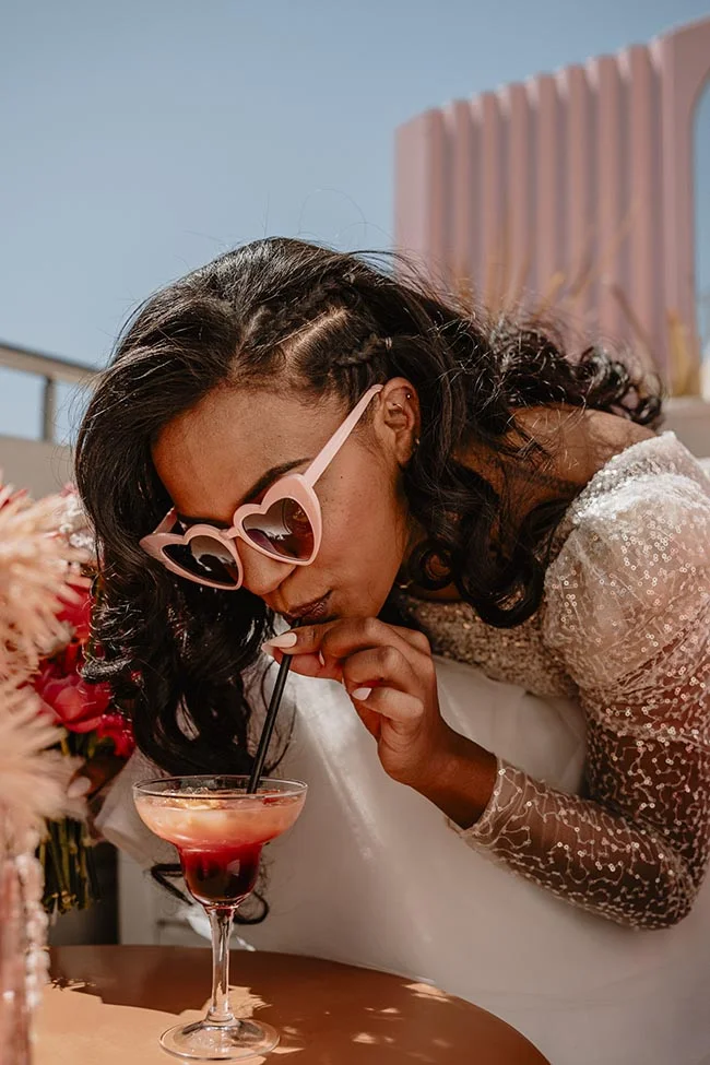 Une mariée rieuse avec des lunettes cœur boit un cocktail coloré lors d'un elopement ensoleillé à Nice.