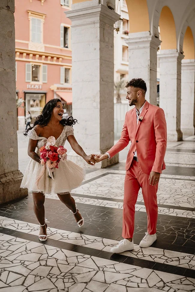 Couple joyeux main dans la main à Nice, elle en robe courte blanche et lui en costume corail, sous des arcades.