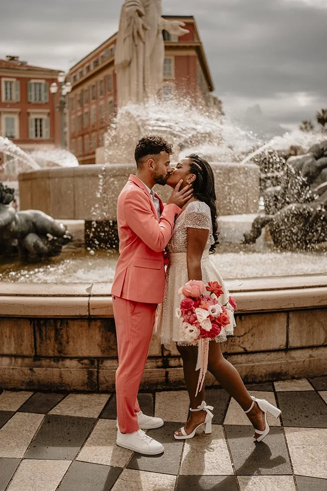 Couple s'embrassant devant une fontaine à Nice, l'homme en costume corail et la femme en robe blanche courte.