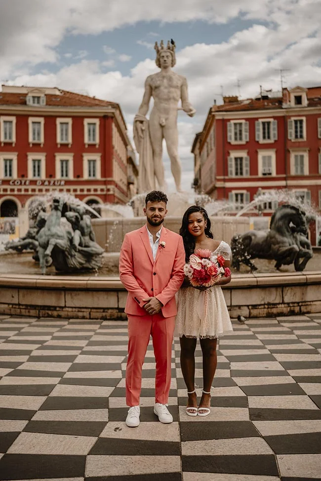 Couple élégant posant devant la fontaine du Soleil à Nice, l'homme en costume corail et la femme en robe blanche.
