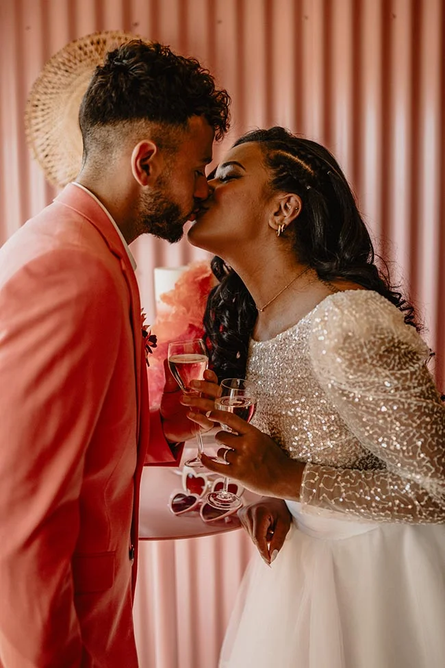Couple s'embrassant avec complicité, elle en robe pailletée et lui en costume corail, devant un fond rose poudré.