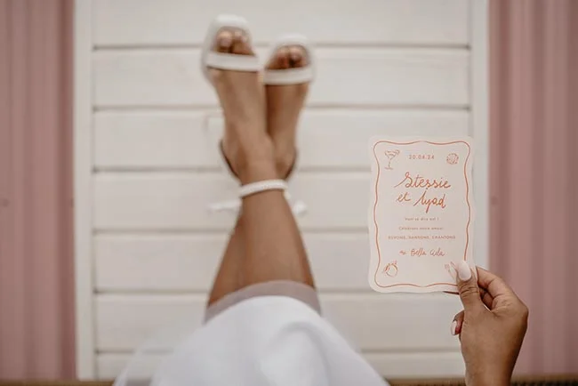 Jambes levées d'une mariée en peignoir blanc tenant un faire-part minimaliste dans un décor aux tons poudrés.