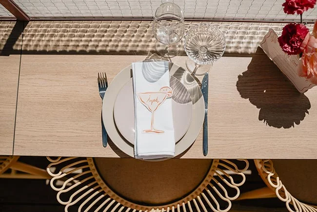 Dressage de table minimaliste et chic avec une serviette blanche illustrée d'un cocktail sur une table en bois clair.