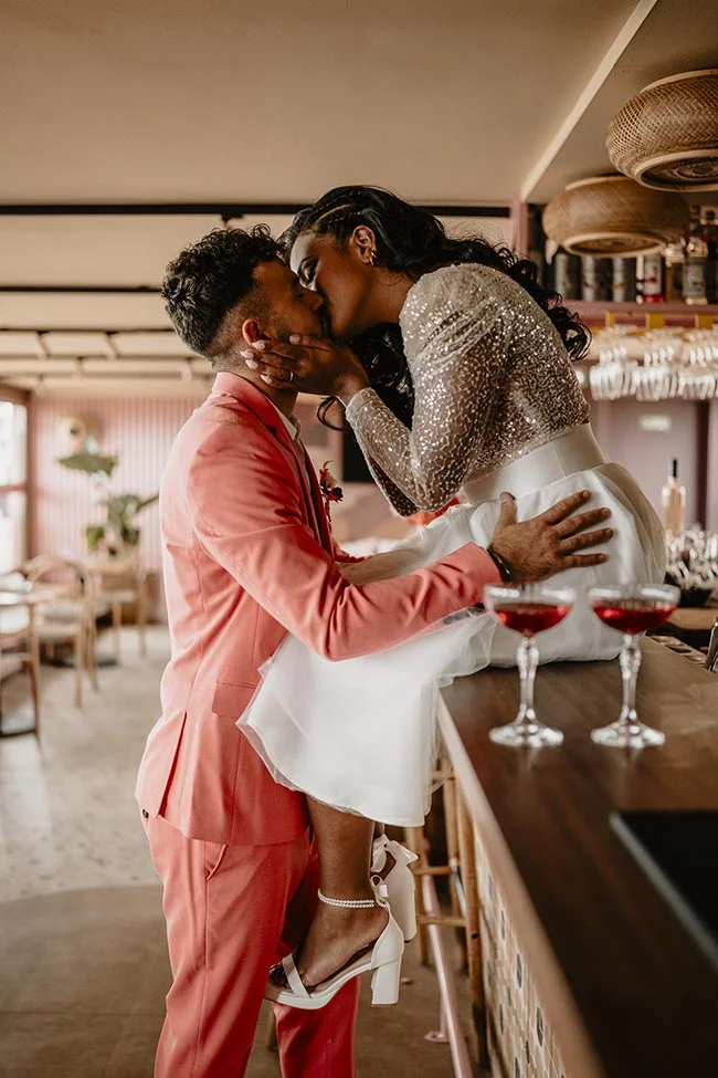 Couple s'embrassant avec passion, elle en robe pailletée et lui en costume corail, dans un bar chic et lumineux.