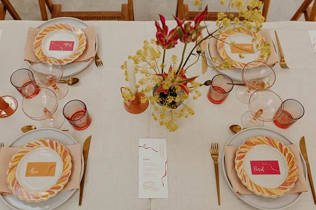 Table dressée pour un mariage coloré avec vaisselle jaune et rose, verres teintés et compositions florales exotiques.