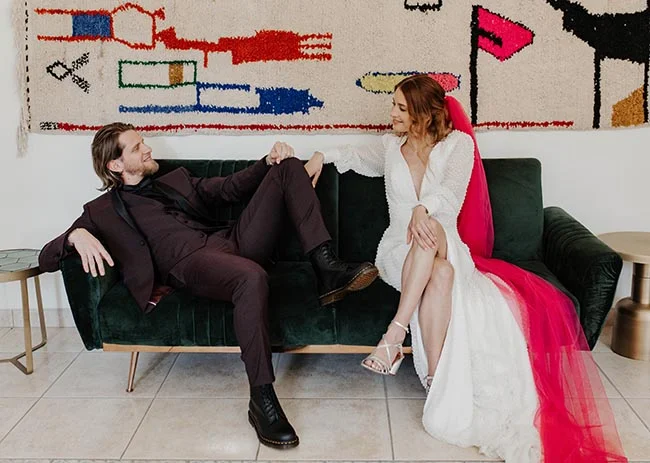 Couple marié décontracté sur un canapé vert devant une œuvre colorée, elle porte une robe blanche et un voile rose vif.