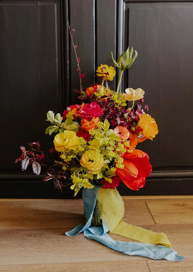 Bouquet floral éclatant aux tons rouge, orange et jaune, noué de rubans bleu et jaune sur un sol en bois.
