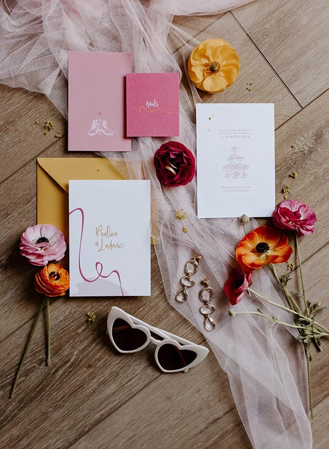 Papeterie de mariage colorée, fleurs éclatantes et lunettes cœur sur un fond bois avec un voile vaporeux.