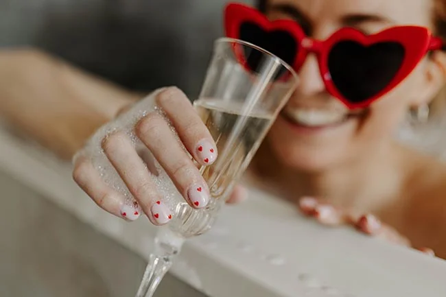 Femme souriante avec lunettes en forme de cœur et ongles décorés, tenant une flûte de champagne dans un bain moussant.