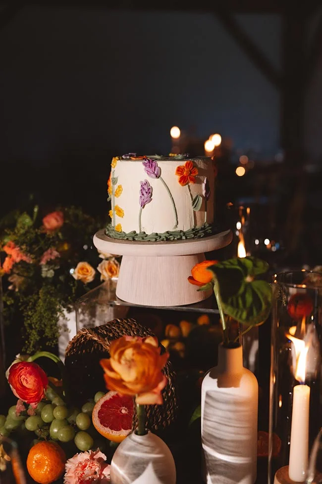 Gâteau de mariage blanc décoré de fleurs colorées sur un support, entouré de fruits et bougies dans une ambiance tamisée.