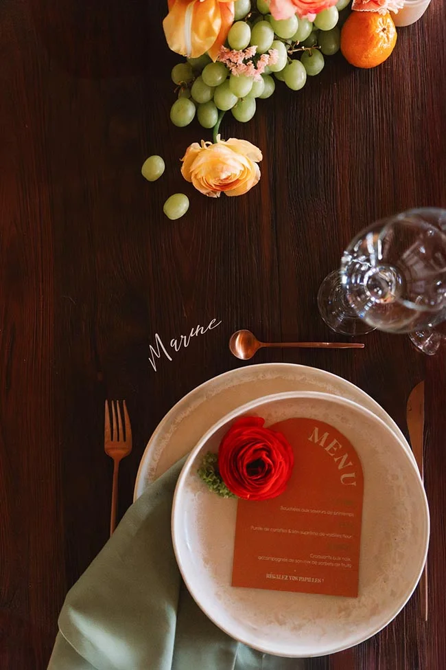 Dressage de table élégant avec assiette, menu terracotta, rose rouge et fruits sur un fond de bois sombre.