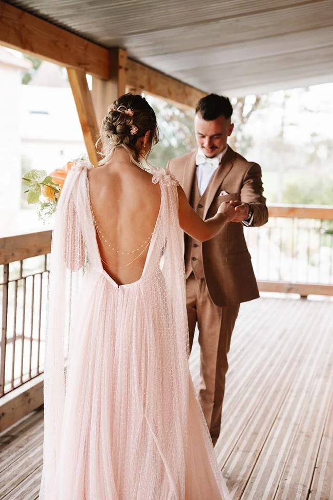 Couple de mariés s'enlaçant sur une terrasse en bois, la mariée en robe rose poudré et le marié en costume marron.