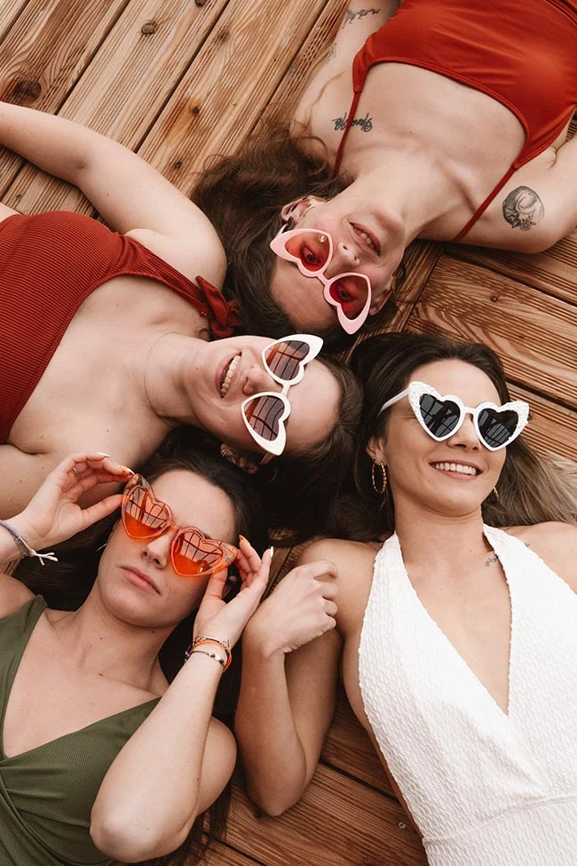 Quatre amies souriantes avec des lunettes en forme de cœur, allongées sur un ponton en bois dans une ambiance festive.