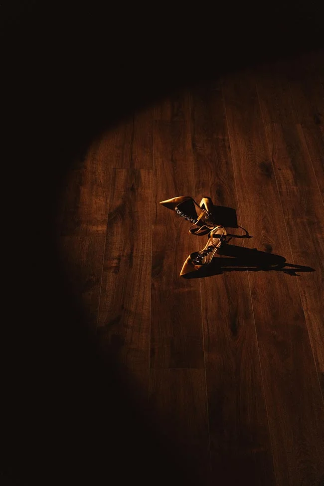 Paire de chaussures à talons dorées posée sur un parquet sombre, éclairée par un faisceau de lumière dramatique.