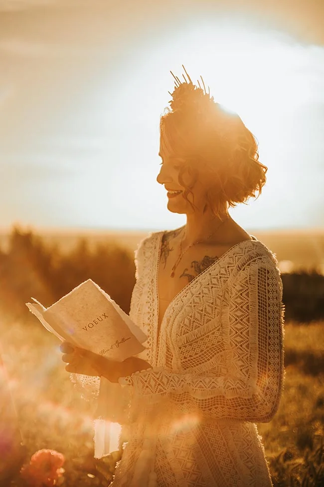 Mariée souriante en robe de dentelle lisant ses vœux dans un champ doré, baignée par une lumière chaude et solaire.
