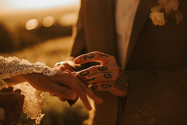 Gros plan chaleureux d'un marié tatoué passant une alliance au doigt de sa mariée sous une lumière dorée de coucher de soleil.