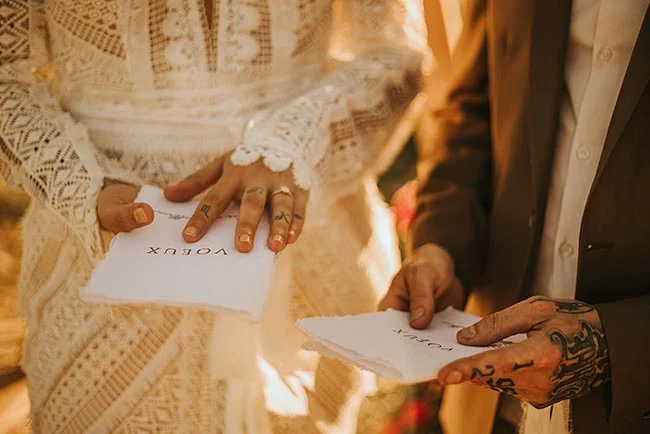 Gros plan d'un couple tenant leurs vœux, ambiance chaleureuse et dorée, robe en dentelle blanche et costume sombre.
