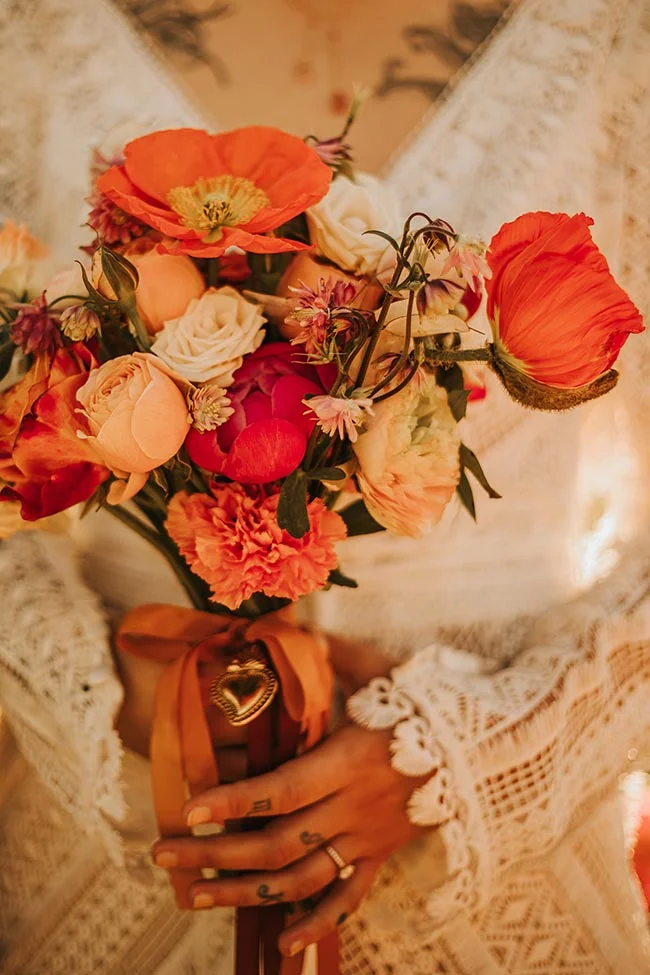 Gros plan d'une mariée tenant un bouquet coloré de fleurs orange et rouges sur une robe en dentelle blanche.