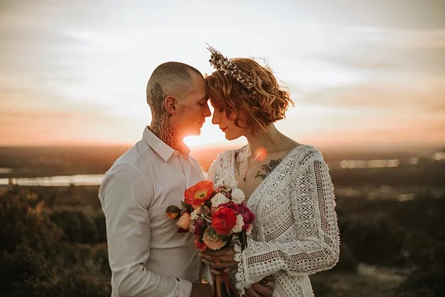 Couple en tenue bohème blanche s'embrassant tendrement au coucher du soleil dans un paysage sauvage et doré.