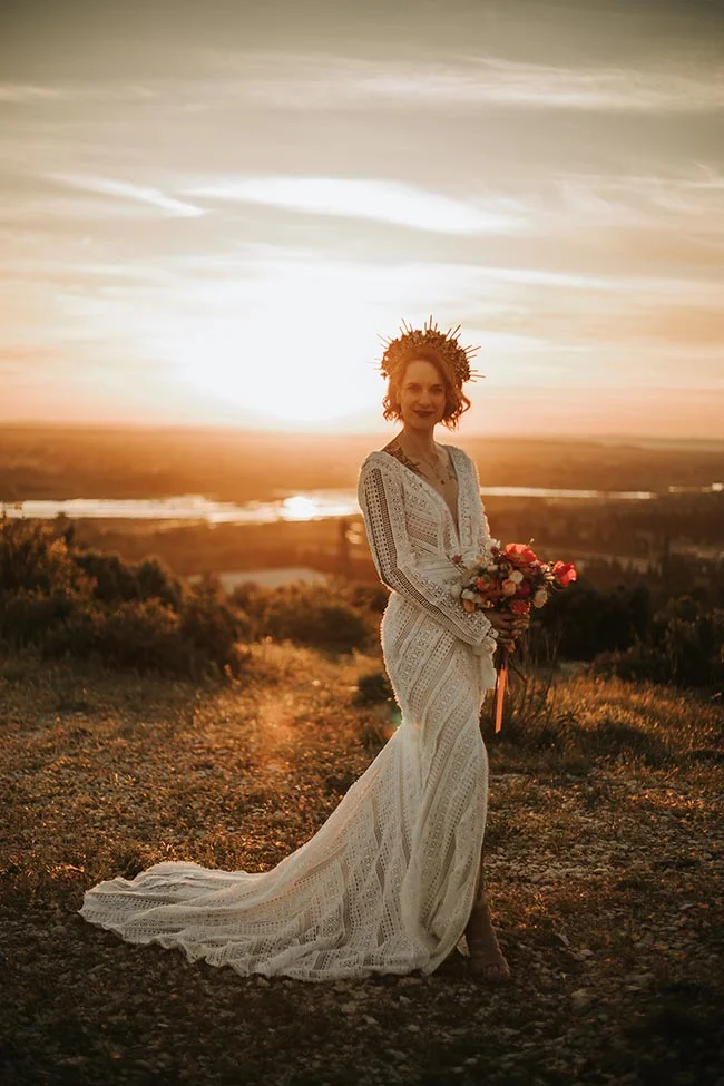 Mariée en robe blanche dentelle et couronne solaire, posant au coucher du soleil dans un paysage sauvage doré.