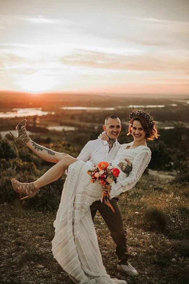 Un couple souriant s'enlace au coucher du soleil dans un paysage sauvage, la mariée portant une robe en dentelle blanche.